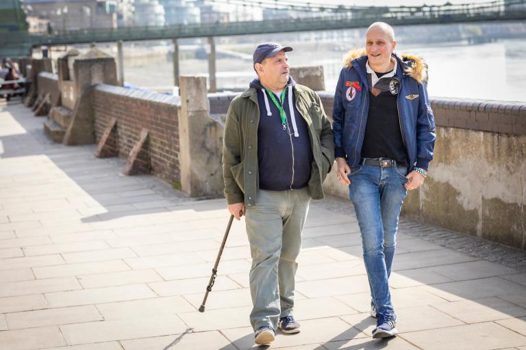 Two men walking near river in London, one has a cane