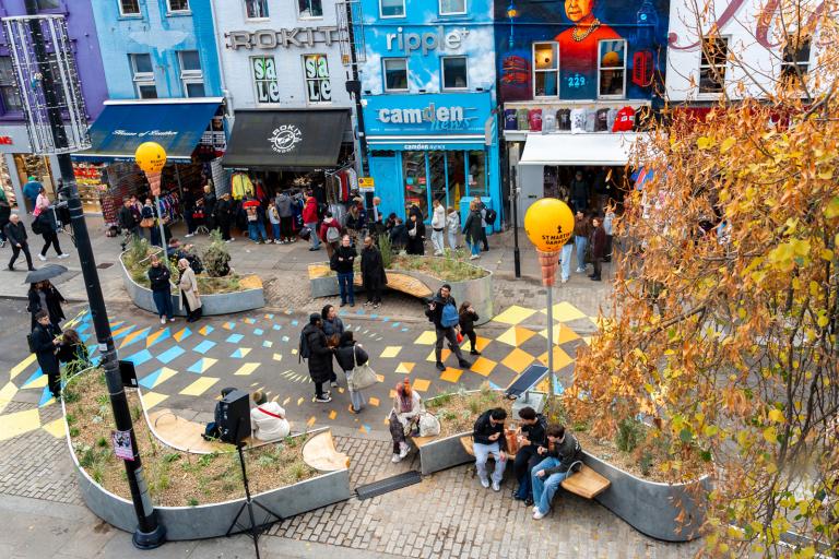 Image shows Camden High Street with colourful shopfronts. Pedestrians are walking along the road, the surface of which has been decorated with colourful shapes. Pedestrians are also sitting on benches next to curved planters.