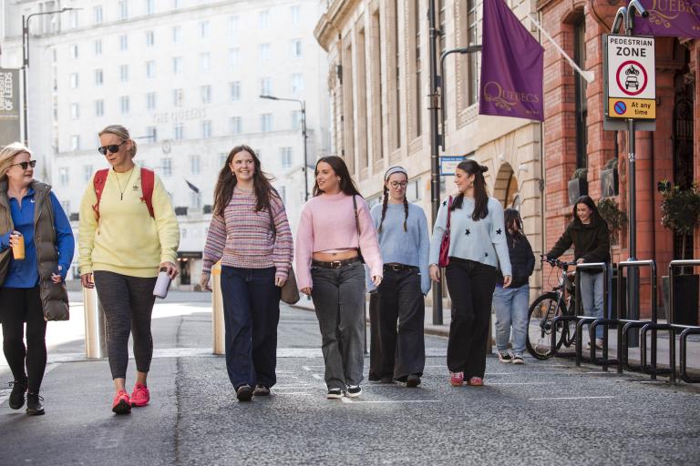 A group of women walk along a pavement in an urban area.