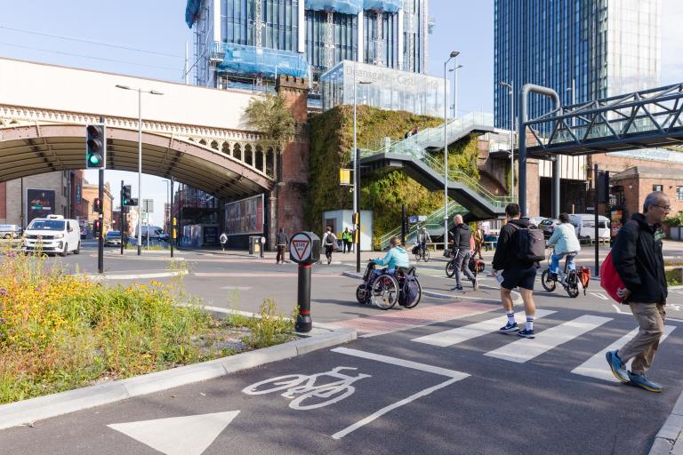 A junction in an urban area. Cyclists are using cycle lane, and pedestrians are crossing a zebra crossing towards walking towards the signalised pedestrian crossing.