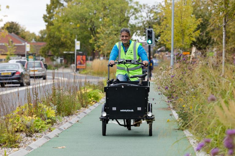 A man wearing a high vis jacket cycles along a cycle track by a road. He is using a cargo bike.