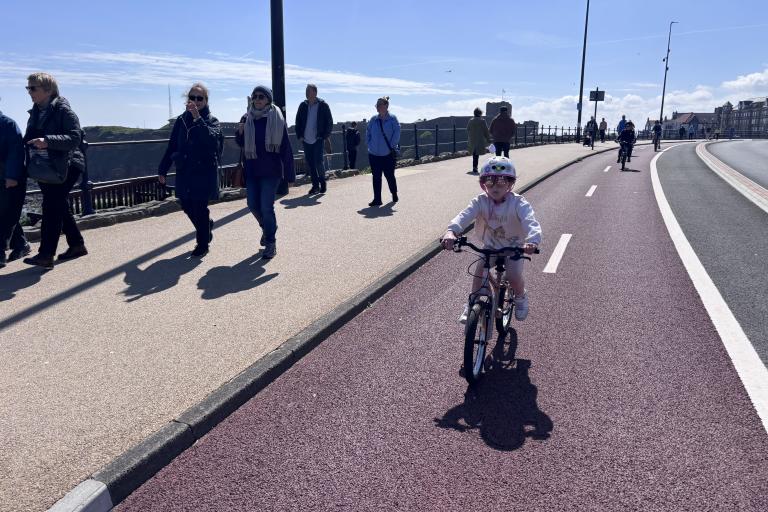 Child cycles along a cycle track separated from the road and the pavement. Pedestrians walk along the pavement.