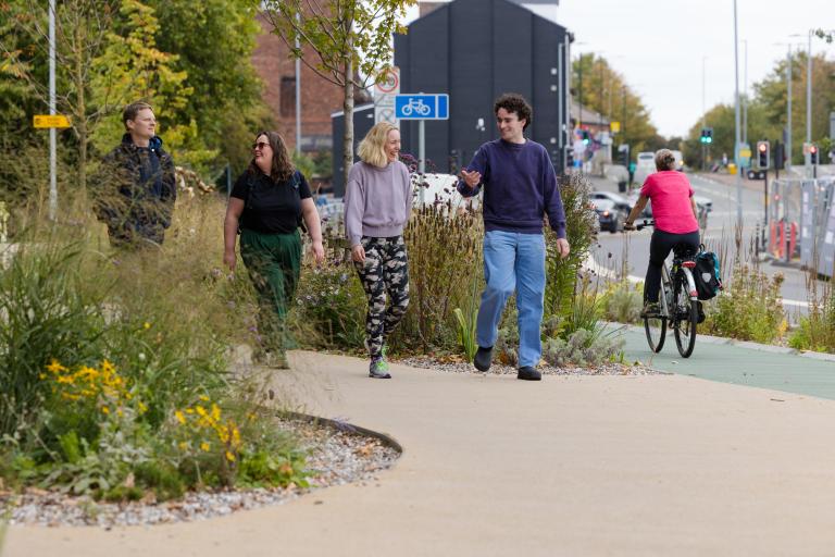 People walking along a surfaced path by a cycle track next to a road. The path is bordered by green vegetation.