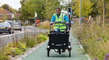 A man wearing a high vis jacket cycles along a cycle track by a road. He is using a cargo bike.