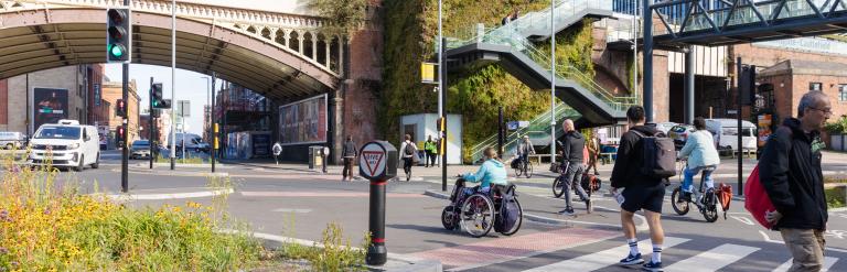 A junction in an urban area. Cyclists are using cycle lane, and pedestrians are crossing a zebra crossing towards walking towards the signalised pedestrian crossing.
